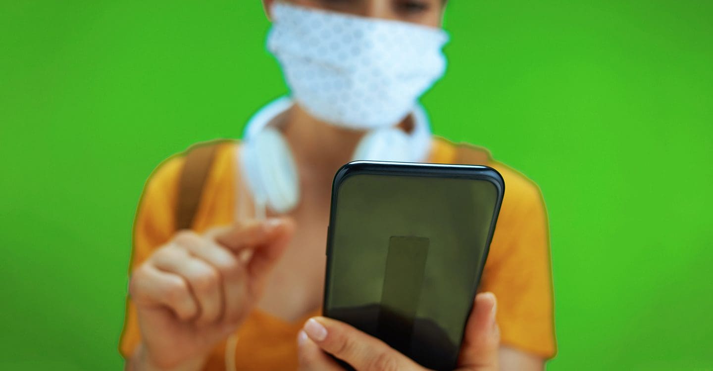 Woman with headphones and wearing a mask typing on her phone with a green background